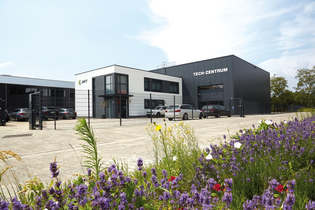 Bild von dem Firmengebäude von JMT CNC Fraesmaschinen in Oberhausen. Blühende Sommerblumen im Vordergrund und das neue moderne Tech-Zentrum von JMT